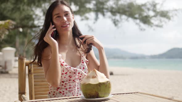 Cheerful Woman with Coconut Talking on Phone on Seaside alt