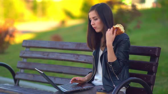 An Attractive Young Girl, Eats a Bun and Works at a Laptop