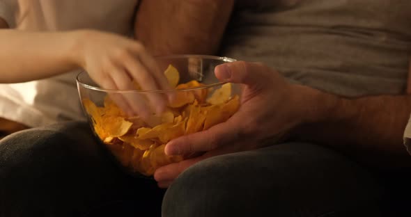 Men's and children's hands take chips from a bowl. Bowl on knees. Close up