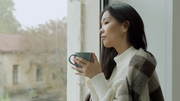 Young Sad Asian Woman Drinks Coffee or Tea and Looks Out the Window Covered with a Blanket alt