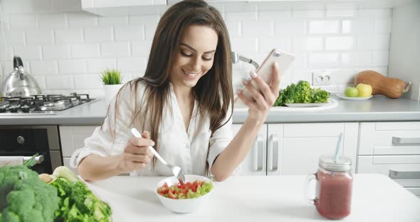 Woman Eating Salad in Kitchen Holding Smartphone