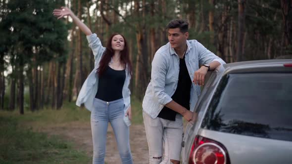 Stressed Handsome Man Leaning on Broken Car with Woman Jumping Gesturing Calling Help alt