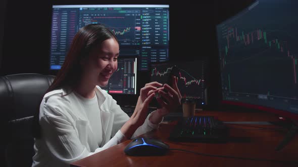 Female Stock Market Broker Using Mobile Phone With Analysing Graphs On Multiple Computer Screens alt