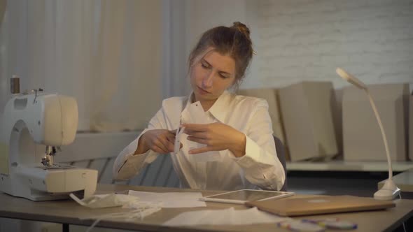 Serious Brunette Tailor Cutting Covid-19 Face Mask with Scissors, Sighing, and Holding Head in Hands alt