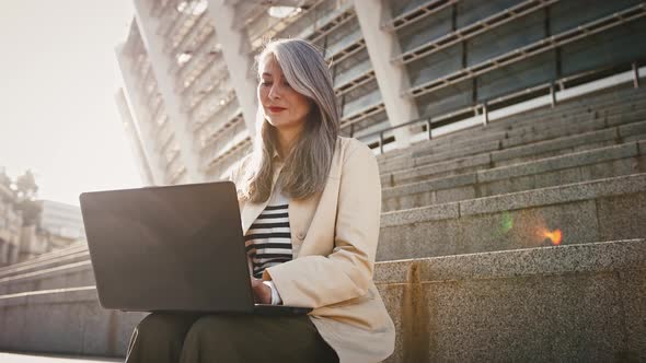 Asian Mature Grayhaired Lady in Casual Outerwear is Working Online at Her Laptop While Sitting on alt