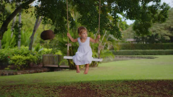 A Little Girl in a White Dress Rides on a Rope Swing in the Park alt