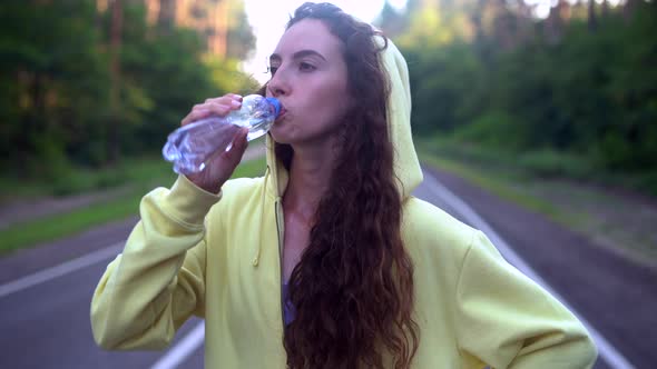 Beautiful Young Caucasian Girl  Standing on Asphalt Forest Road Drinks Water alt