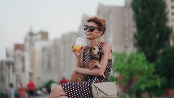 Elderly Woman Resting on Bench on Boardwalk with Crowd of People with Small Dachshund Dog in Her alt