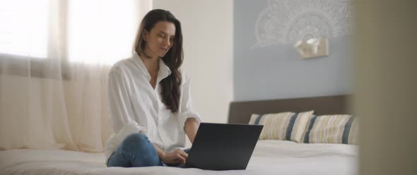 A young woman smiling while working on a laptop from her bedroom alt