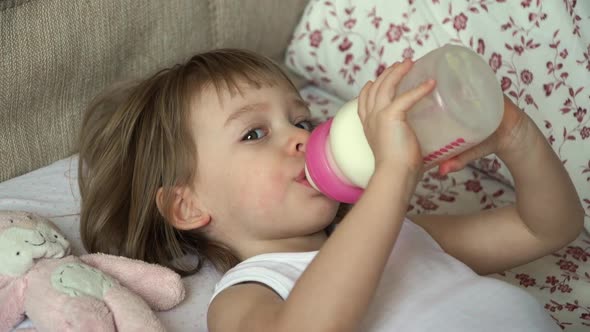 Little Baby Lies on Bed Waking Up in Morning Takes Bottle of Milk and Sucks alt