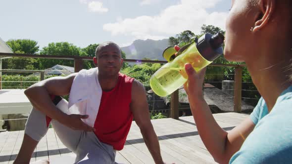 Tired biracial couple on terrace drinking water, resting after exercises and talking alt