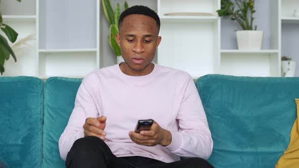 Happy Young African Ethnicity Man Relaxing on Cozy Sofa at Home, Holding Remote Controller, Watching alt