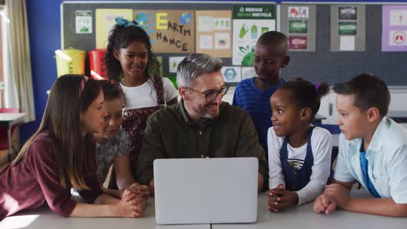 Diverse male teacher and group of schoolchildren looking at laptop alt