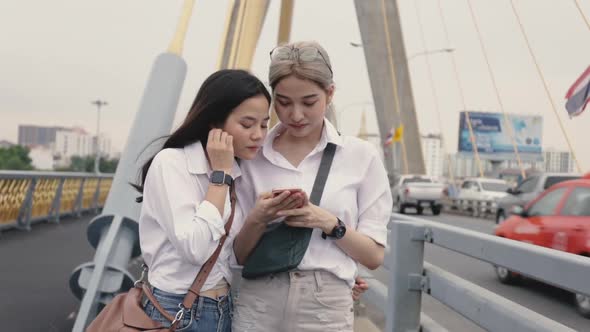 Asian lesbian couples using smartphones browsing the website while standing on the bridge.