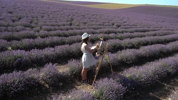 A Woman in a Dress Paints a Picture of a Landscape in a Blooming Lavender Field alt