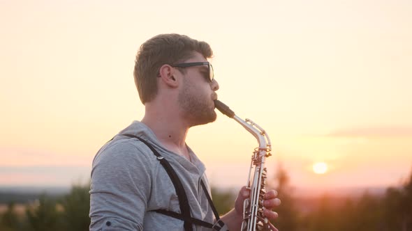 Bearded Man in Glasses and Gray Turtleneck and Suspenders Play Silver Saxophone Outdoors in alt