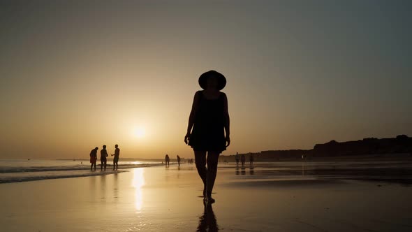 The Silhouette of a Woman in a Summer Dress and Panama Walks Along the Beach at Low Tide at Sunset