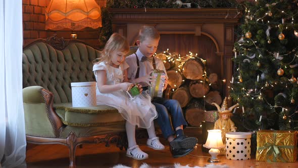 Happy little children sitting on the couch near the Christmas tree opens gifts alt