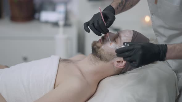 Side View Young Man Lying with Closed Eyes As Tattooed Male Beauty Therapist Applying Moisturizing alt