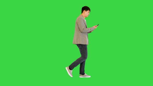 Young Asian Man Walks Looking at His Smartphone on a Green Screen Chroma Key alt