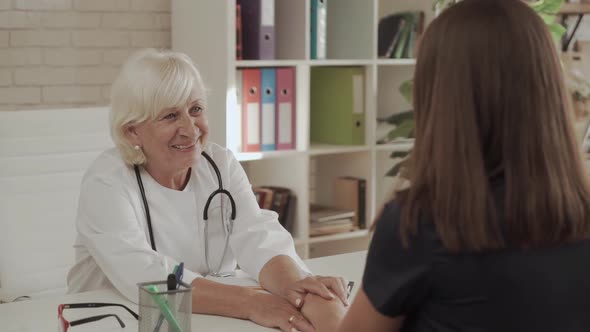 Elderly Woman Doctor Advises Young Woman Patient and Holds Her Hand in the Office alt