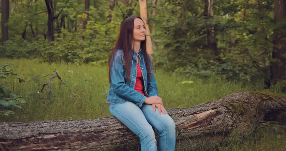Pretty Young Woman Sits on a Log in the Forest and Enjoys Nature