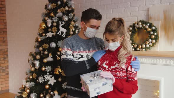 Man Hugging His Girlfriend Give Gift on Xmas Wearing Medical Masks and Gloves alt