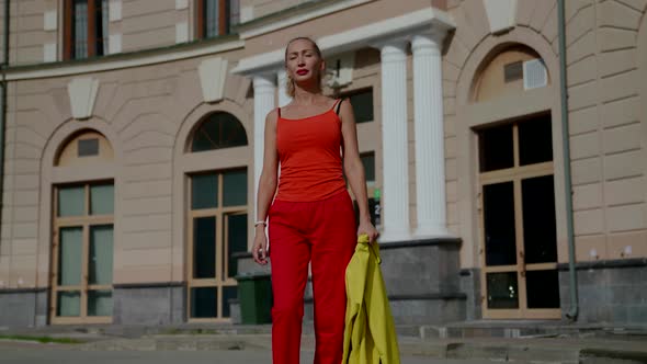 a Woman in Red Clothes Holds a Yellow Jacket in Hand and Walks Against the Background of a House alt