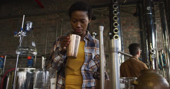 African american woman working at gin distillery smelling product and using tablet alt