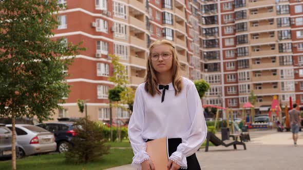 A Schoolgirl Girl with a Notebook in Her Hands Walks Through the Courtyard alt