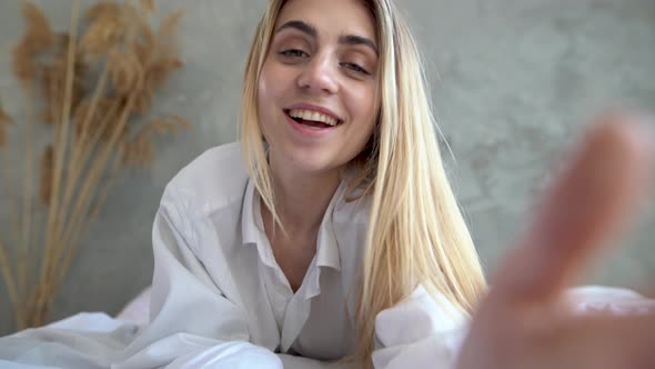 Portrait of Happy Young Caucasian Girl in Casual Clothing Lying Down on Bed While Making Video Call alt
