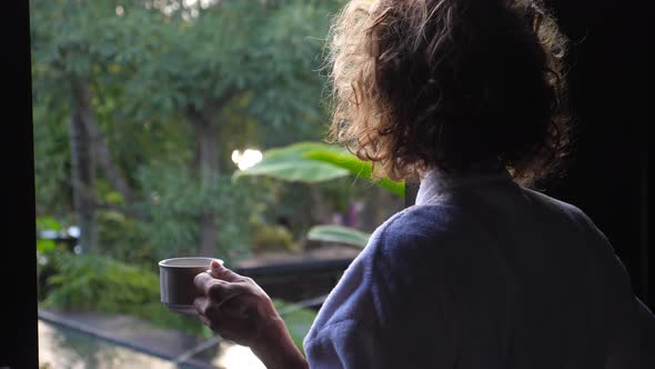 Back Shot of Woman Opening a Window Holding Cup of Coffee alt