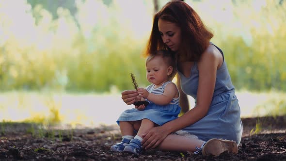 mother and baby sit in a park or in a forest on the ground looking at twigs and cones alt