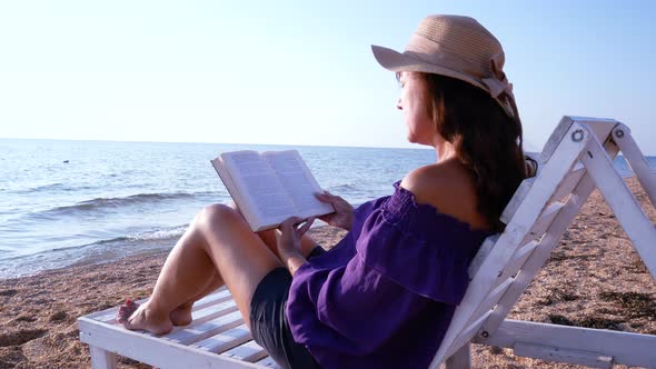 Girl Sitting on a Sun Lounger on the Beach and Reading a Book