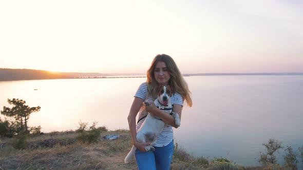 Young Attractive Woman Playing with a Dog Jack Russell in the Meadow at Sunset with Sea Background alt