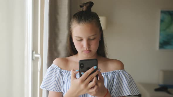A Teenage Girl Standing at the Window and Use a Smartphone alt