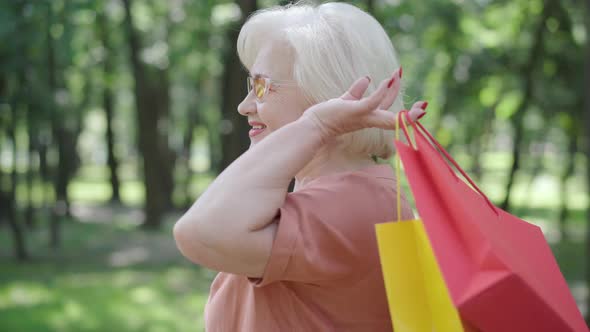 Side View of Happy Elegant Retiree Standing with Shopping Bags Outdoors and Smiling. Portrait of alt