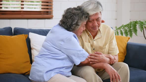 Asian senior elderly couple sitting on a sofa, woman giving hugging to man, support, understanding. alt