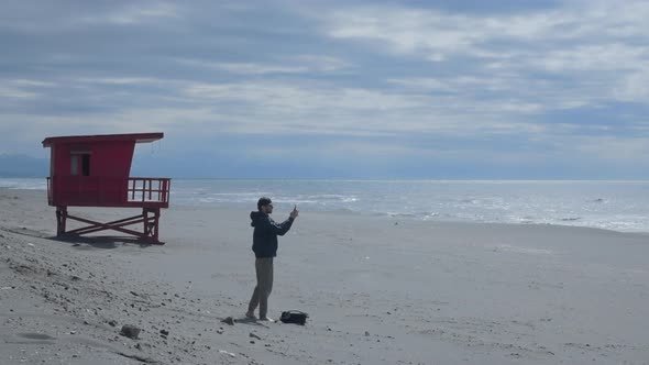Young Male Blogger at Beach with Lifeguard Tower Shoots Story on Mobile Phone