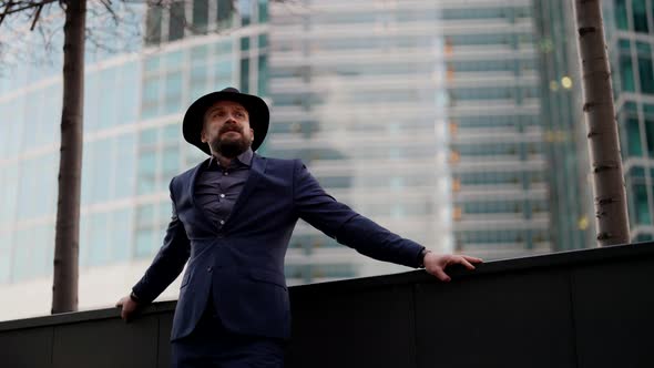 a Bearded and Mustachioed Stylish Man in a Plaid Suit Leaned Hands on the Parapet and Then Takes Off alt