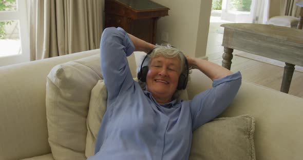 Caucasian senior woman wearing headphones smiling while listening to music at home alt