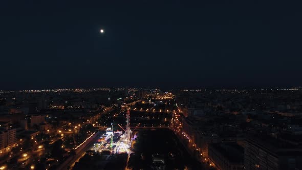 Aerial night view of lighted ferris wheel in amusement park against sky with moon, Valencia, Spain alt
