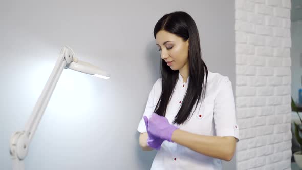 A cute cosmetologist in a white lab coat stands near the wall and disinfects gloves alt