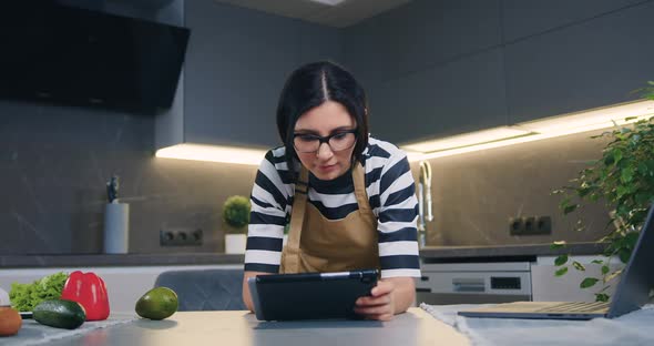 Woman in Glasses Leaning on Kitchen Table and Browsing Apps on Tablet PC alt
