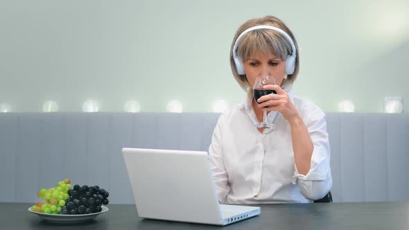 A Beautiful Elderly Woman Sitting at a Laptop Drinking Red Wine From a Glass Studying and Typing