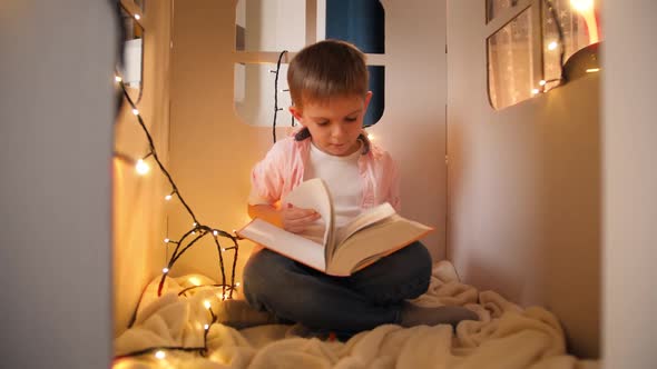 Cute Little Boy Sitting in Toy Cardboard House at Night and Reading Big Fairy Tale Book alt