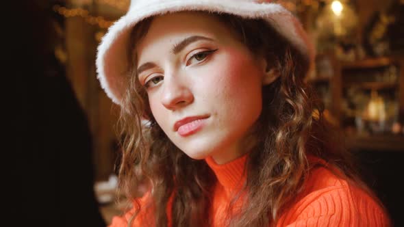 Attractive Beautiful Girl in a Cafe Looks Cute. Young Curly Green-eyed Woman in a Stylish Fluffy Hat