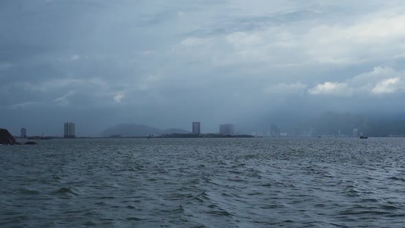 View From Middle of Wavy Sea, Dark Clouds Over a Hazy City and Mountains. alt