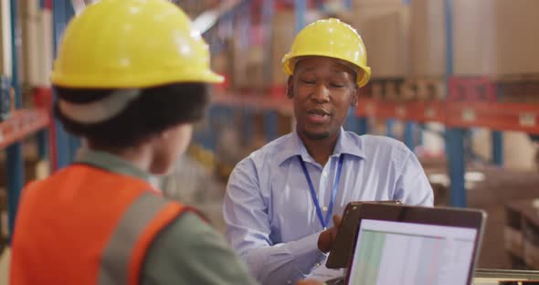 African american male and female workers wearing helmet and using laptop in warehouse alt