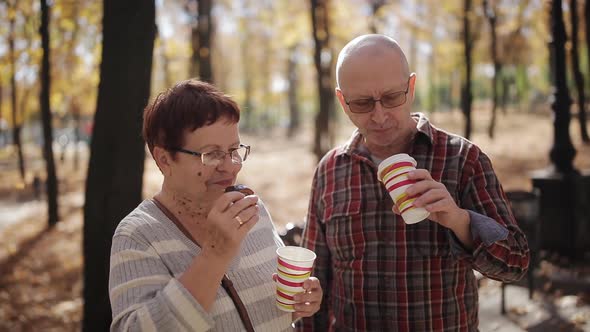 An Elderly Couple in the Autumn Park Talking and Drinking Coffee or Tea in Plastic Cups